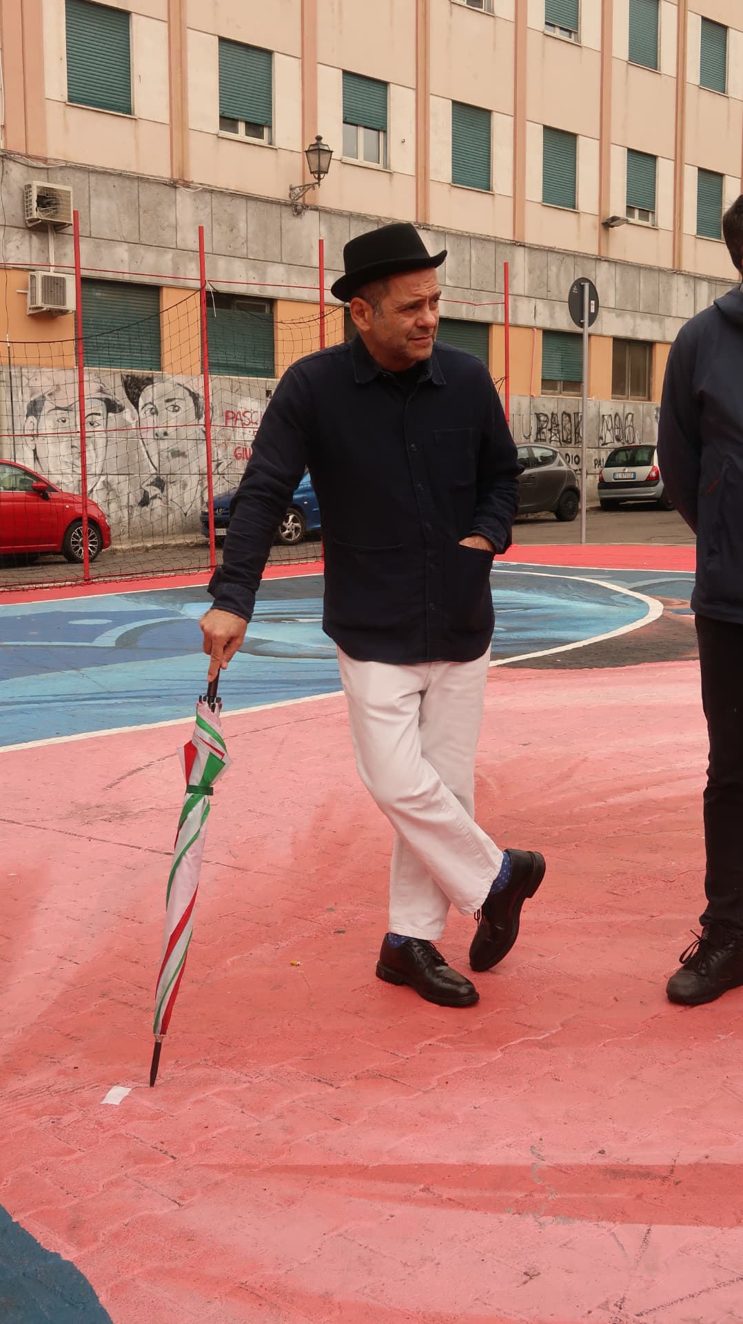 Man in a bowler hat with a folded umbrella crossing a painted court