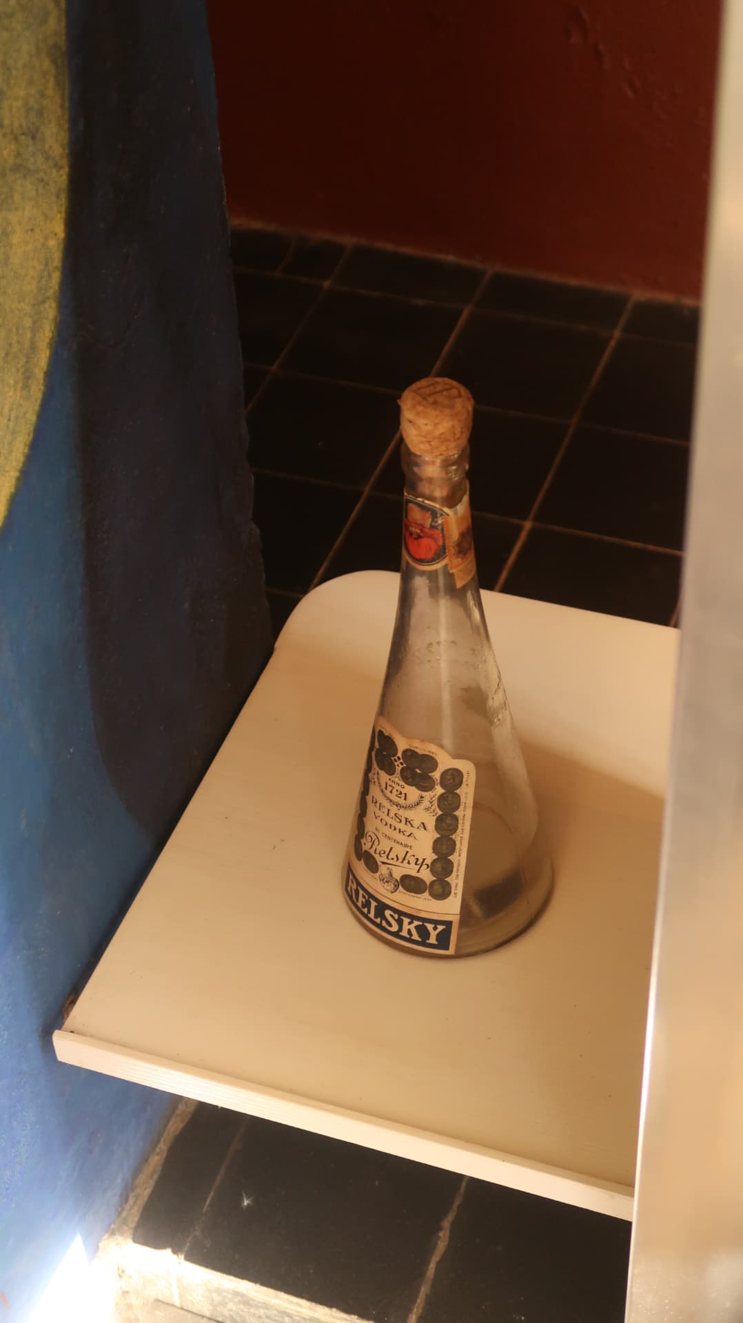 Old vodka bottle with a cork on a white shelf by blue plaster