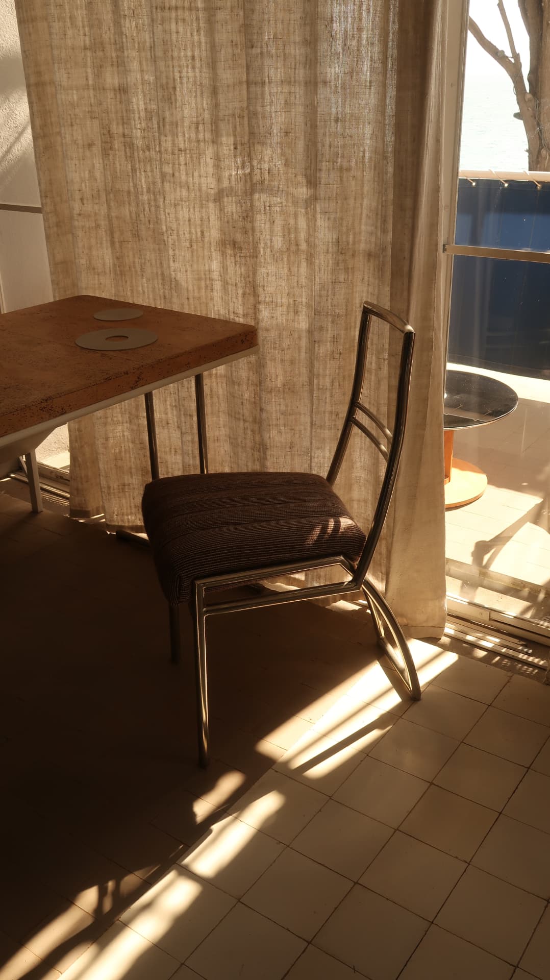 Cork table and chrome chair in raking light behind a linen curtain