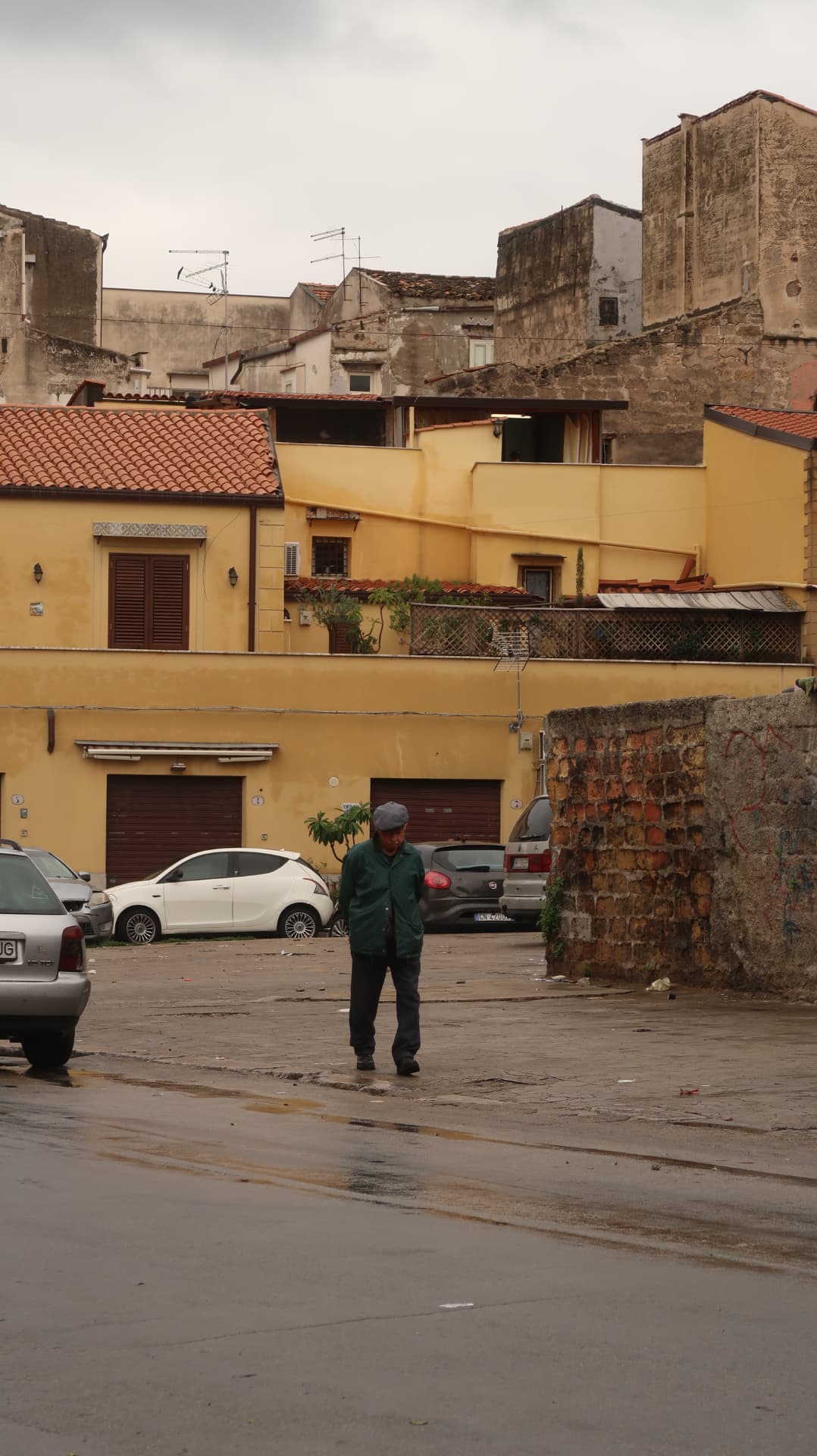 Man in a flat cap crossing an empty yard below ochre houses
