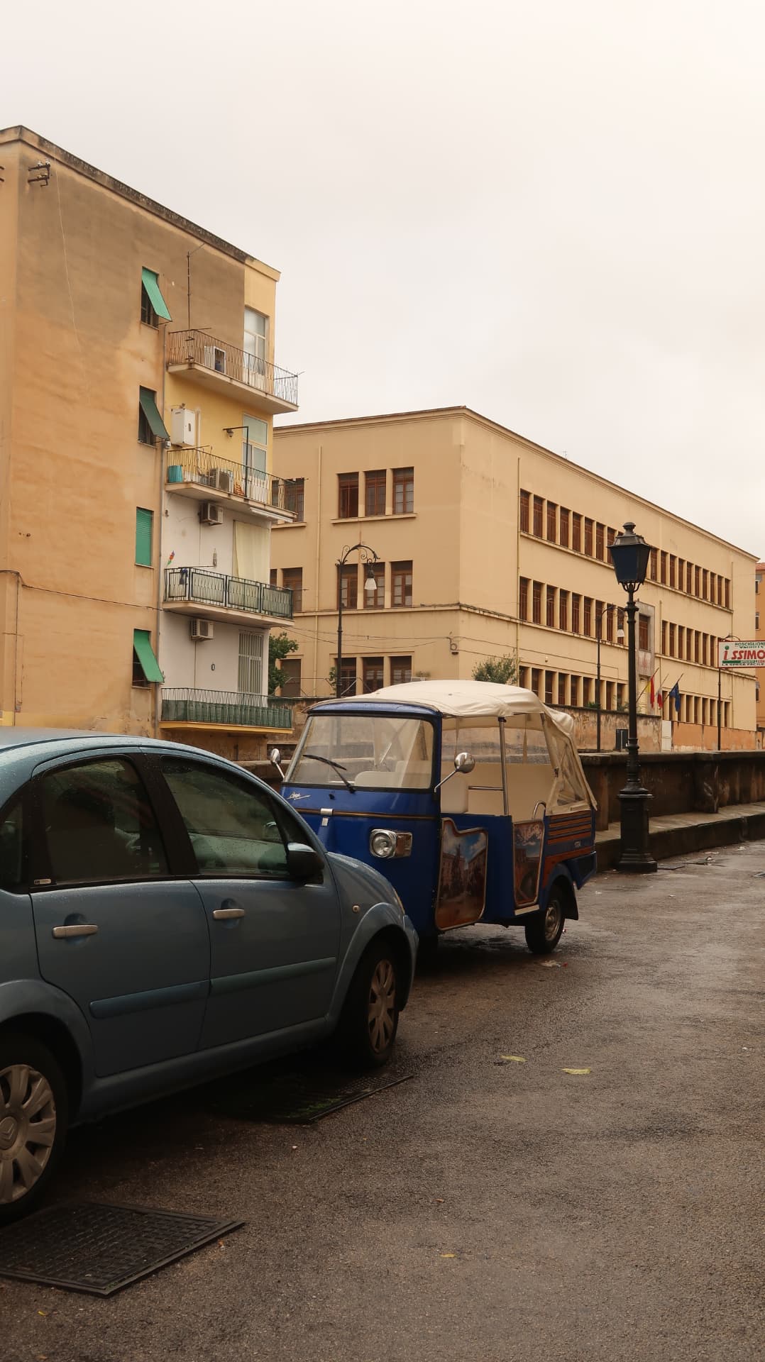Blue three wheeled Ape van parked beside a car on a wet street