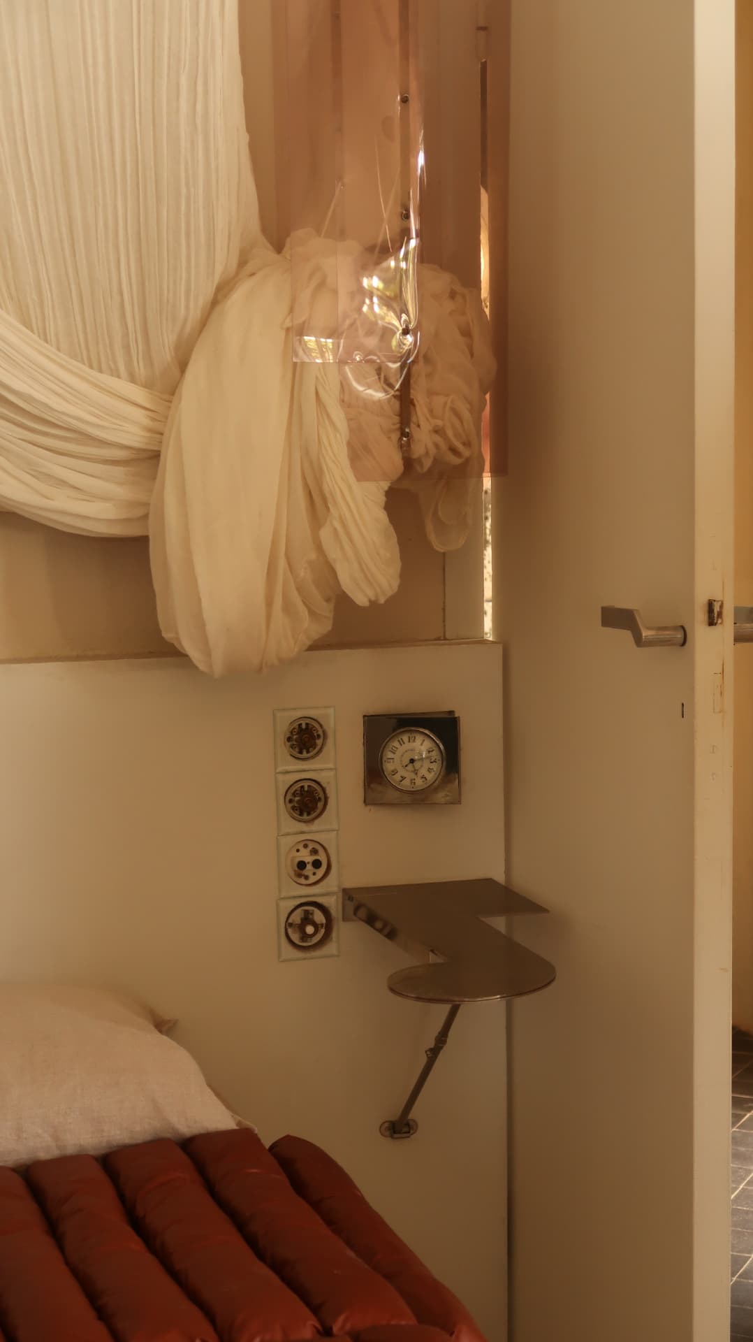 Muslin drapery above a folding steel shelf and a red mattress