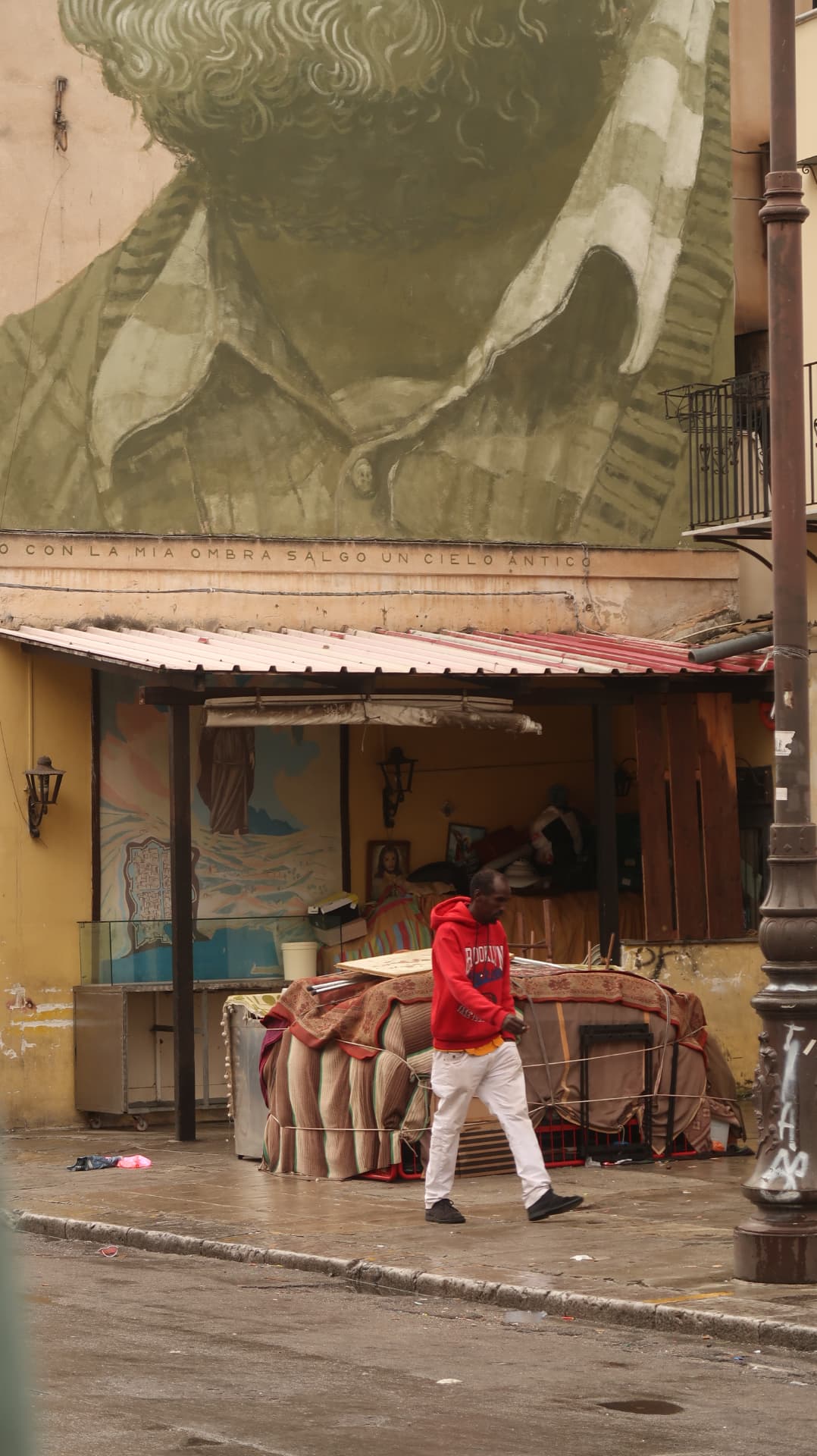 Man in red walking beneath a large green mural and market stall