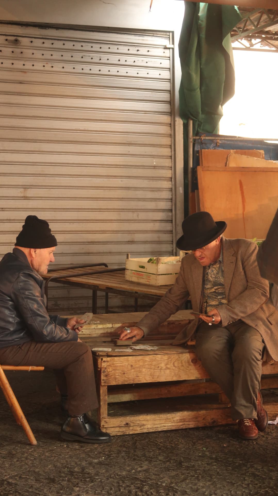 Two men playing cards on a wooden crate in a market shed