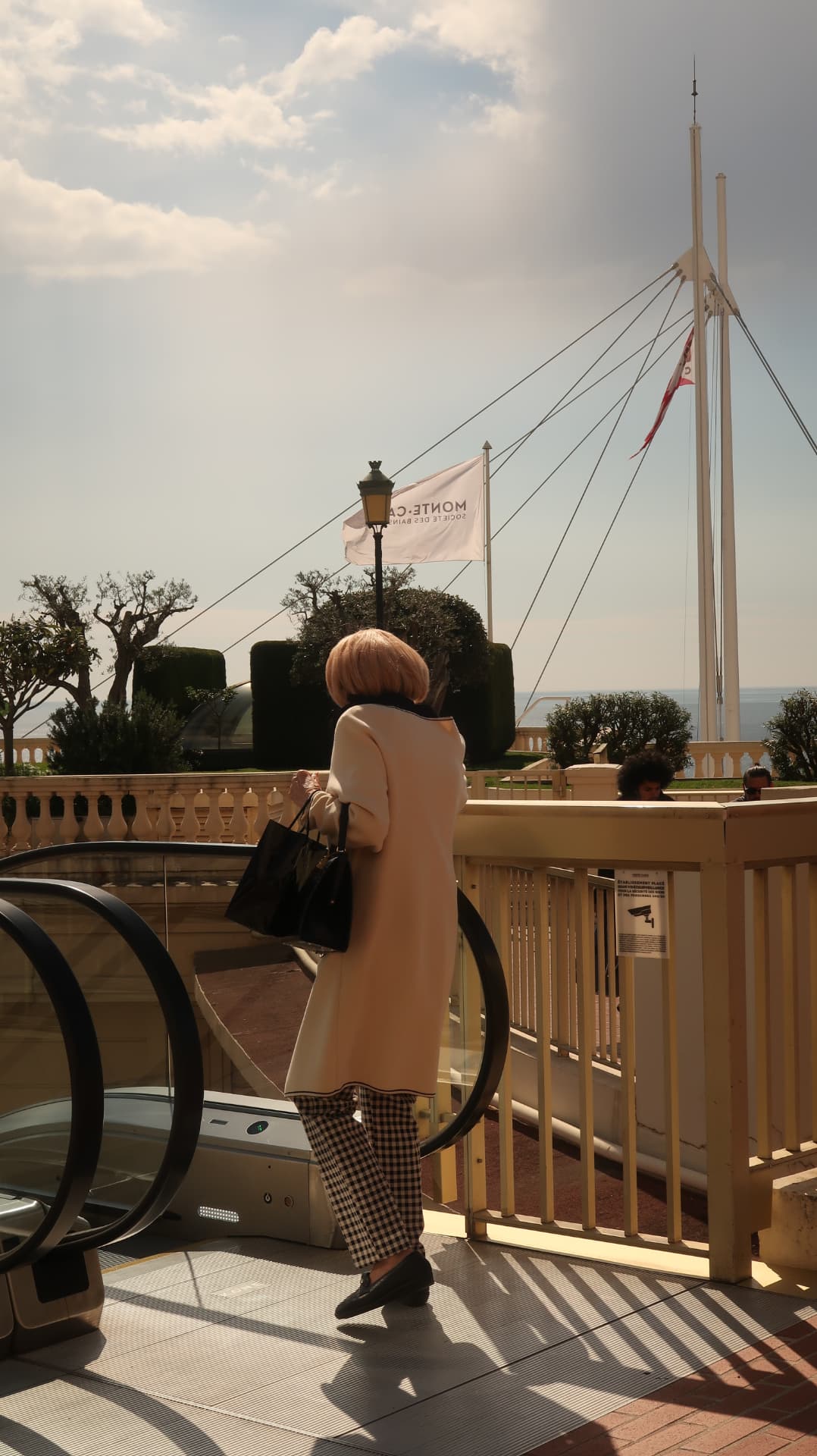 Woman in a cream coat at the top of an escalator by the sea