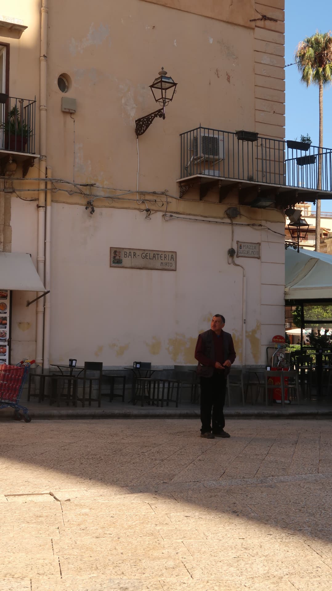 Man standing in a piazza below a mosaic Bar-Gelateria sign