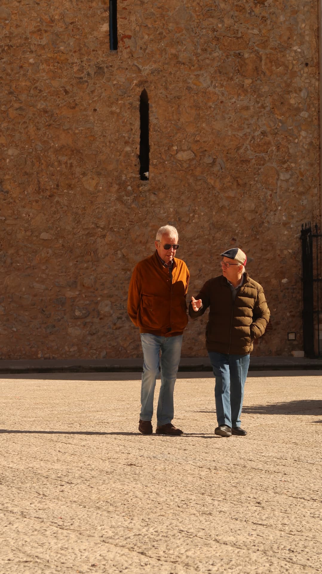 Two men walking and talking across a sunlit square before a rough stone wall