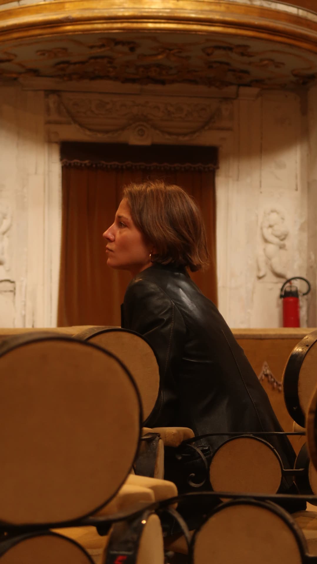 Woman in a leather jacket seated in profile before a gilded balcony