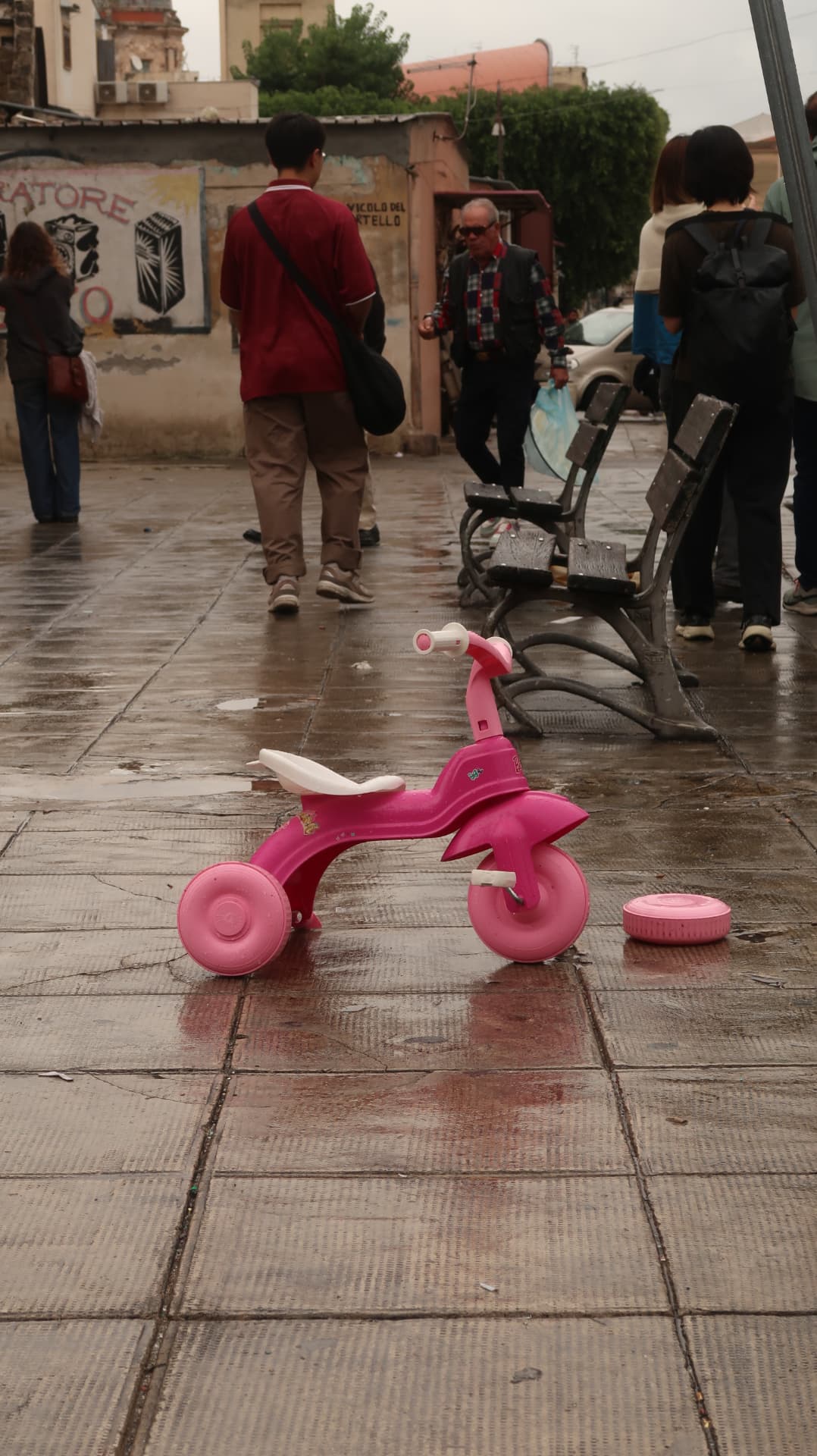 Pink toy tricycle abandoned on wet paving in a busy square