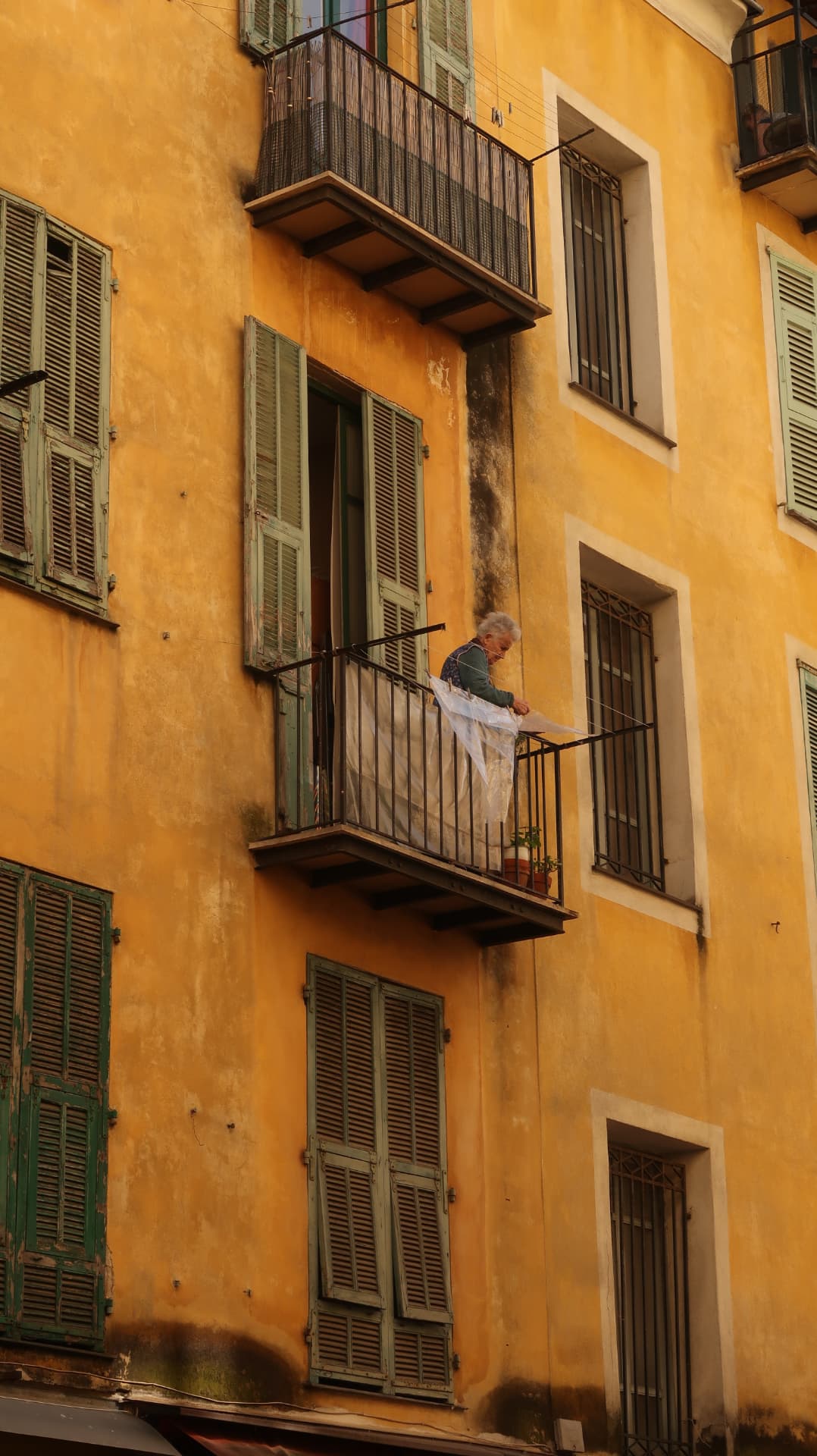 Woman shaking out linen on a balcony of an ochre facade